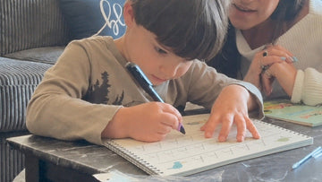 A young boy tracing a letter inside a grooved copybook page while an adult points at the row beside him.