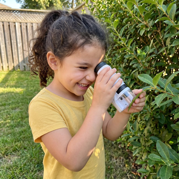 A smiling child holds the microscope to one eye, with its clear base against a leaf on a green hedge