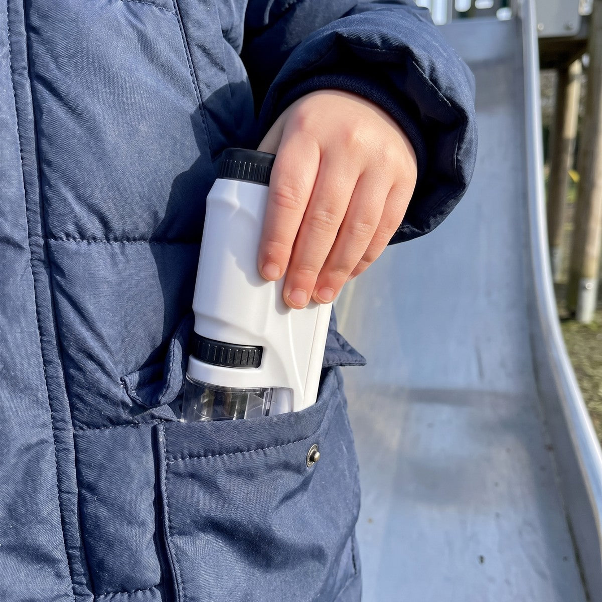 A child slides the microscope into the front pocket of a puffy jacket at a playground, showing it is pocket-sized