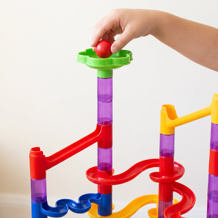 A child's hand drops a red ball into a green funnel at the top of a clear purple tube